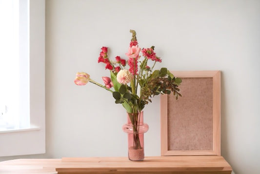 Kunstbloemen Veldboeket Paars/ Roze/ Pink inclusief Glasvaas op een houten tafel met een lichte muur met raam en fotolijstje