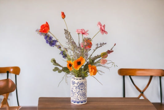 Kunstbloemen Veldboeket Lavende/ Roze/ Oranje inclusief Vaas met oa papaver Cosmea en Ridderspoor op een bruine houten tafel met een witte muur en stoelen