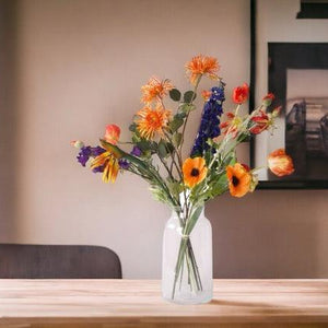 Kunstbloemen Veldboeket Rood/ Blauw/ Oranje inclusief Glasvaas in de huiskamer op een houten tafel voor een bruine muur met poster op de achtergrond