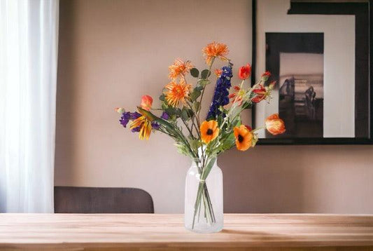 Kunstbloemen Veldboeket Rood/ Blauw/ Oranje inclusief Glasvaas in de huiskamer op een houten tafel voor een bruine muur met poster op de achtergrond