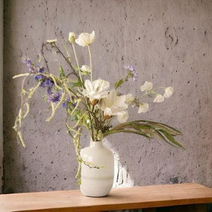Kunstbloemen Veldboeket wit met blauw inclusief vaas op een bruine houten tafel voor het raam tegen een grijze muur