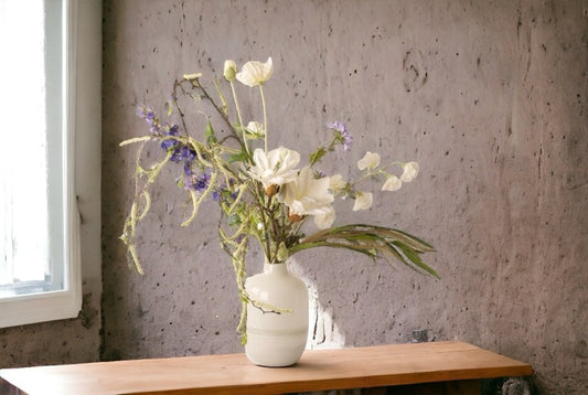 Kunstbloemen Veldboeket wit met blauw inclusief vaas op een bruine houten tafel voor het raam tegen een grijze muur