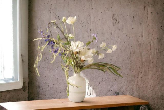 Kunstbloemen Veldboeket wit met blauw inclusief vaas op een bruine houten tafel voor het raam tegen een grijze muur