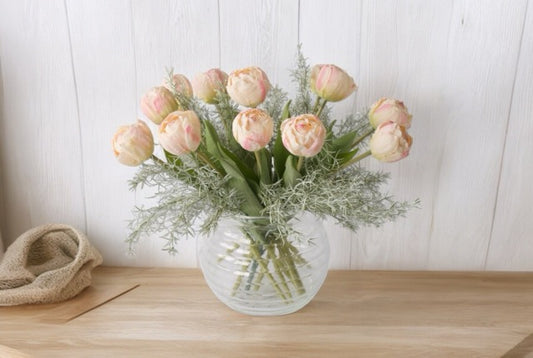 boeket van roze tulpen met bladgroen in een bolle glasvaas op een houten tafel voor een witte Mur