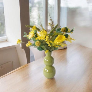 Veldboeket Zijden bloemen in de kleur Geel op een houten tafel in een groene hoogglans metalen vaas op een houten tafel voor een raam