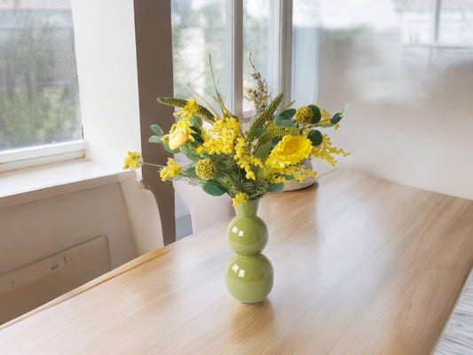 Veldboeket Zijden bloemen in de kleur Geel op een houten tafel in een groene hoogglans metalen vaas op een houten tafel voor een raam