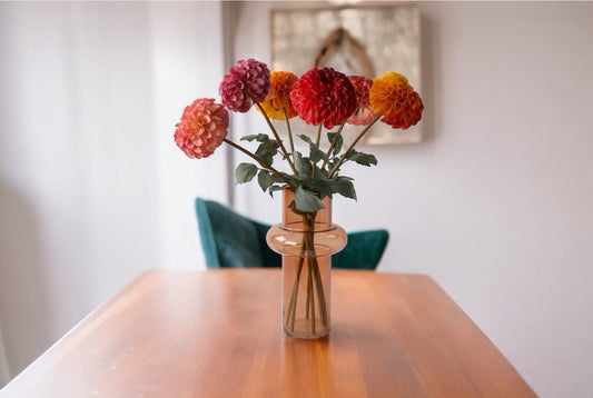 Boeket zijden dahlia's in de kleuren rood oranje mauve en pink in licht roze glas vaas op een houten tafel