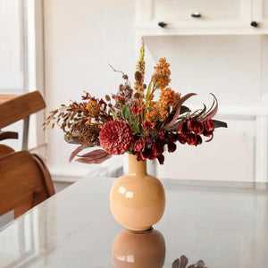 Boeket met kunstbloemen op een beige kleurige vaas  in bordeaux rode en oranje kleuren bestaande uit Physalis, Magnolia en Chasmantium op een glazen tafel in een keuken