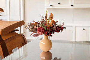 Boeket met kunstbloemen op een beige kleurige vaas  in bordeaux rode en oranje kleuren bestaande uit Physalis, Magnolia en Chasmantium op een glazen tafel in een keuken