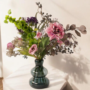 Boeket kunstbloemen in lila , grijs en paars kleuren op grote blauwe glasvaas op een tafel voor een lichte muur en houten vloer