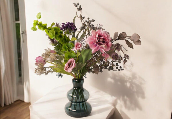 Boeket kunstbloemen in lila , grijs en paars kleuren op grote blauwe glasvaas op een tafel voor een lichte muur en houten vloer