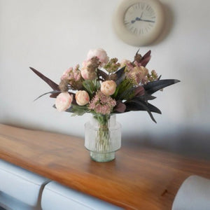 Kort Gebonden Boeket Kunstbloemen Zacht Rozebos Tulp zacht roze Fluitekruid Roze kastanje Stone Ranoinkel Roze Tack Eucalyptus Grijs/ Rood op een glasvaas van helder glas op een houten tafel voor een lichte muur met een klok - WinQ