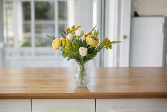 Fris Voorjaars Kunstbloemen Boeket in Geel met Wit met Tulpen wit, Fluitekruid geel, Ranonkel Geel, Tak Mimosa op een glas vaas van helder glas op een tafel van hout voor een raam