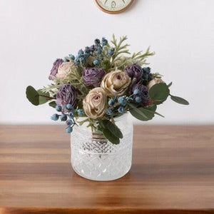 Kortgebonden Kunstboeket Rozen in lila en zacht lila op een glasvaas hoogte van het kunstbloemen boeket is 23cm op een houten tafel met een klok op de achtergrond