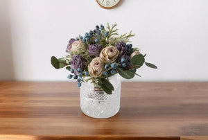 Kortgebonden Kunstboeket Rozen in lila en zacht lila op een glasvaas hoogte van het kunstbloemen boeket is 23cm op een houten tafel met een klok op de achtergrond