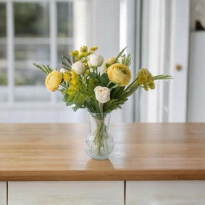 Fris Voorjaars Kunstbloemen Boeket in Geel met Wit met Tulpen wit, Fluitekruid geel, Ranonkel Geel, Tak Mimosa op een glas vaas van helder glas op een tafel van hout voor een raam