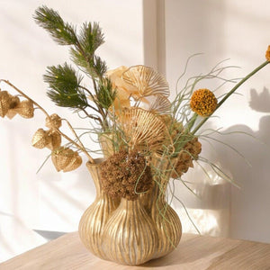 Tulpenvaas met 5 gaten en boeket met kunstbloemen in goud en oker waaronder Physalis en Ginko op een lichte houten tafel voor een lichte muur