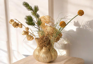 Tulpenvaas met 5 gaten en boeket met kunstbloemen in goud en oker waaronder Physalis en Ginko op een lichte houten tafel voor een lichte muur