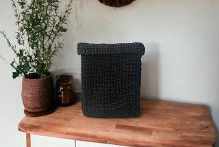 Opbergmand Abaca Vierkant 25x25x35cm Zwart op een houten tafel voor een lichte muur  en wat planten