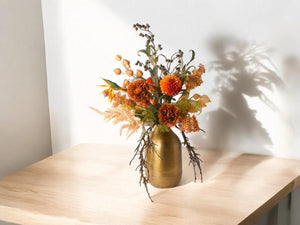 Boeket kunstbloemen in diverse kleuren crème, oranje,terra op een koperkleurige vaas op een houten tafel voor een lichte muur