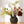 Kunstbloemen in een zwarte tulpenvaas met 3 openingen gevuld met diverse kunstbloemen op een lichte houten tafel voor een witte muur