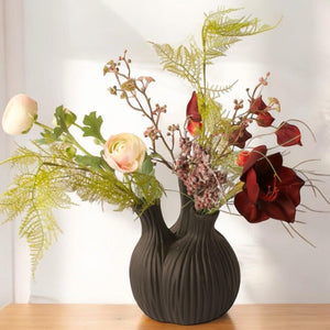 Kunstbloemen in een zwarte tulpenvaas met 3 openingen gevuld met diverse kunstbloemen op een lichte houten tafel voor een witte muur