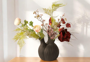 Kunstbloemen in een zwarte tulpenvaas met 3 openingen gevuld met diverse kunstbloemen op een lichte houten tafel voor een witte muur