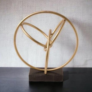 Ornament va goud / brons, opgebouwd uit diverse rondingen en bogen gemaakt van ronde buis, geheel staat op een zwart marmeren voet maat van het geheel is 27x20x30cm geheel staat op een zwarte tafel tegen een witte muur