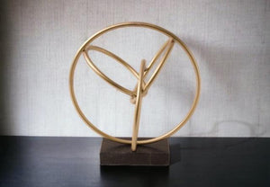 Ornament va goud / brons, opgebouwd uit diverse rondingen en bogen gemaakt van ronde buis, geheel staat op een zwart marmeren voet maat van het geheel is 27x20x30cm geheel staat op een zwarte tafel tegen een witte muur