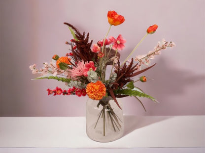 Boeket kunstbloemen in  rood en oranje tinten op een glasvaas met diverse bloemen, waaronder papaver dahlia, scabiosa en ranonkel