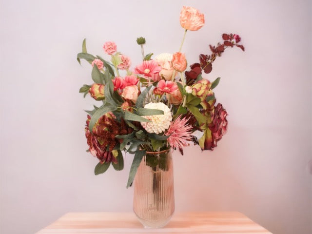Veldboeket met nepbloemen waaronder papaver en pioen in rood en roze combinatie met accenten van groen blad in een roze glasvaas met een hoogte van 65cm op een houten tafel