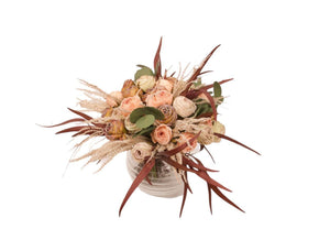 Kort gebonden boeket van kunstbloemen bestaande uit rozen in roze, mauve en rood gepresenteerd op een ronde glasvaas