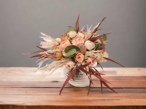 Kort gebonden boeket van kunstbloemen bestaande uit rozen in roze, mauve en rood gepresenteerd op een ronde glasvaas op een houten tafel