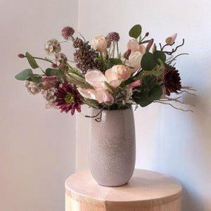 Gebonden kunstbloemen op een keramieken mauve kleurige vaas Het boeket is in een paars Roze kleurencombinatie met onder andere tulpen en gerbera en eucalyptus Boket staat op een houten zuil voor een lichte muur