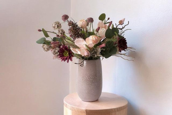 Gebonden kunstbloemen op een keramieken mauve kleurige vaas Het boeket is in een paars Roze kleurencombinatie met onder andere tulpen en gerbera en eucalyptus Boket staat op een houten zuil voor een lichte muur