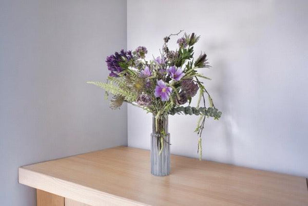 PlukBoeket Kunstbloemen Lila en Blauw op een tafel in een hoek tegen een lichte muur,.