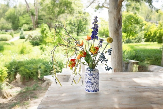 Plukboeket van Zijden Bloemen Blauw, Geel en Oranje Betula Geel Gloriosa Bes Blauw Papaver Oranje Allium Geel en een bijpassende vaas in delfts blauw van keramiek op een tuintafel in de tuin met bomen en struiken op de achtergrond