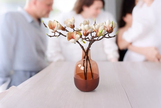 Kunstbloemen Magnolia 3 takken zacht roze inclusief vaas cognac op een tafel met een familie op de achtergrond