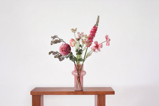 Boeket kunstbloemen in de kleuren roze, pink en rooide een roze glasvaas op een houten tafel tje voor een lichte muur
