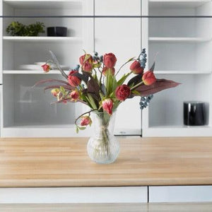 Gebonden Moederdag Boeket in Rood met Hibiscus, Tulp Rood, Bes Blauw, Tak Eucalyptus Grijs/Rood in een glasvaas van helder glas op een keukentafel voor de kastenwand