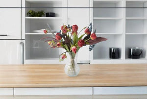 Gebonden Moederdag Boeket in Rood met Hibiscus, Tulp Rood, Bes Blauw, Tak Eucalyptus Grijs/Rood in een glasvaas van helder glas op een keukentafel voor de kastenwand