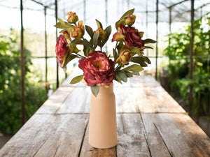 Sfeerfoto van een vaas met 3 takken kunst pioenrozen met meerder bloemen in de kleur bordeaux rood gefotografeerd in een serre op een houten tafel- WInQ