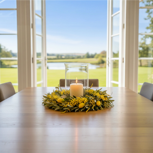 Voorjaarskrans van olijftakken opgemaakt met gele kunstbloemen op een tafel met een windlicht en een kaars in het midden - WinQ