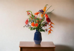 VELDboeket van kunstbloemen in een rood en oranje arrangement op een blauwe hoogglans metalen vaas op een houten kastje voor een lichte muur